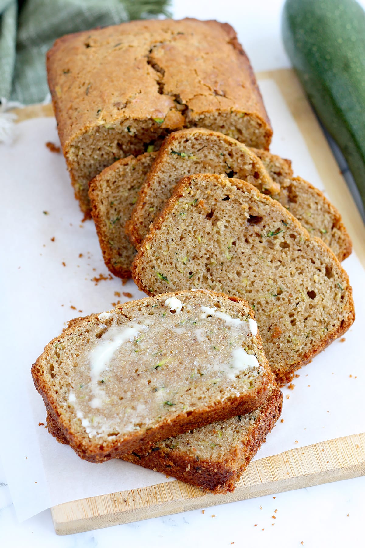 Zucchini bread, sliced and topped with softened butter with zucchini in the background.